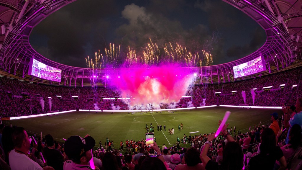 Un estadio lleno de espectadores observando cómo unos vibrantes fuegos artificiales de color rosa iluminan el cielo nocturno.